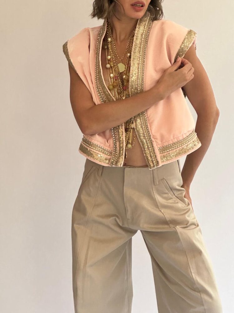 Gilet court en velours rose poudré bordé de galons dorés, porté ouvert sur des sautoirs dorés et un pantalon large beige